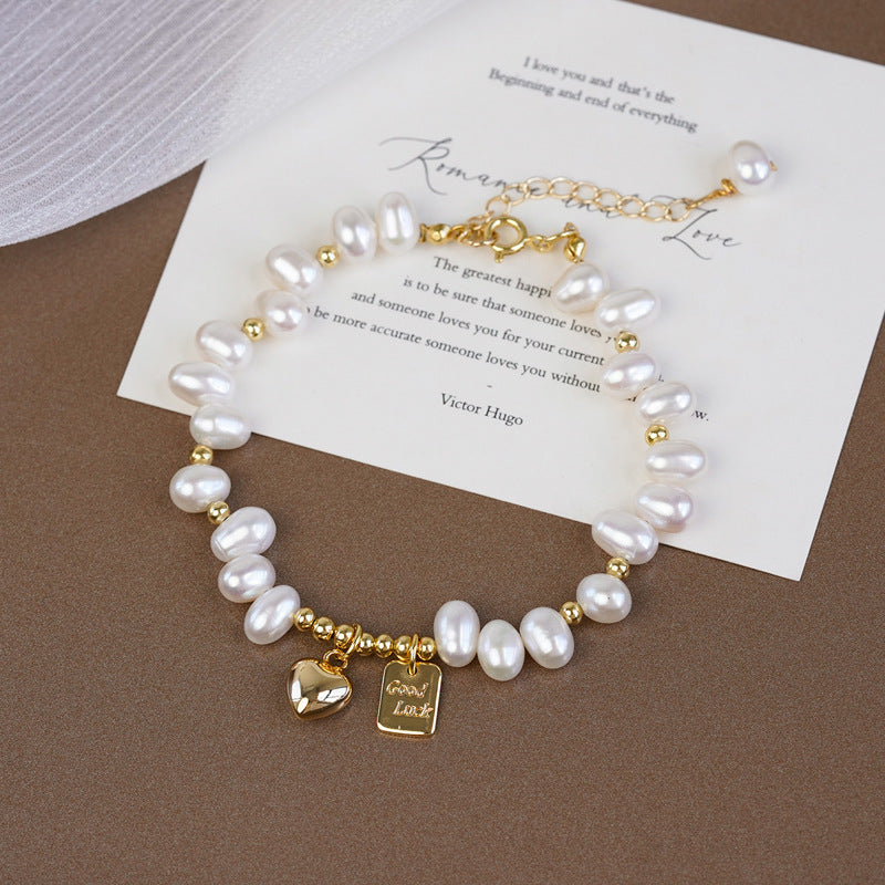 A freshwater pearl bracelet with gold-tone chain and charms, displayed on a brown textured surface over a white card.