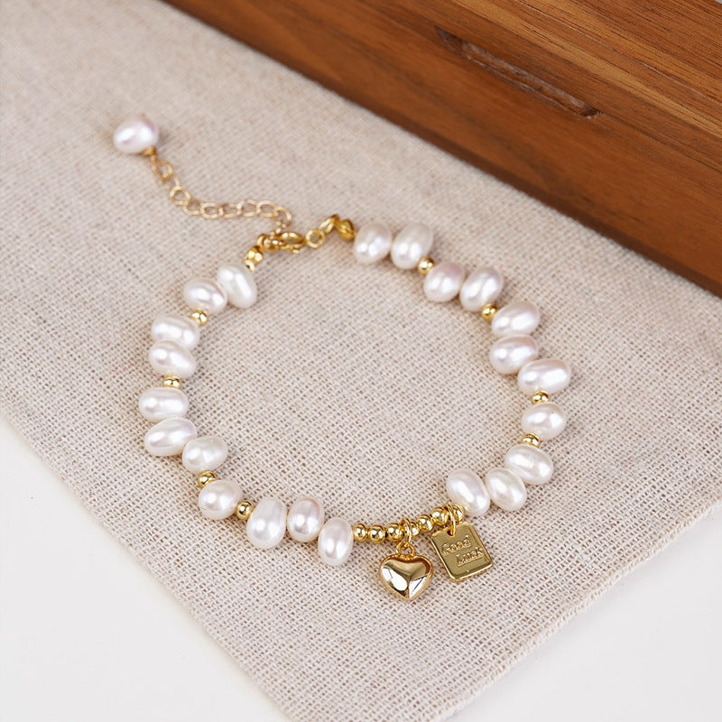 A gold-plated bracelet with irregular white freshwater pearls and two small charms (a heart and a square engraved charm), displayed on a textured beige fabric next to a wooden surface.