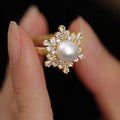 A gold ring with a central white pearl and surrounding clear baguette and round stones, shown worn on a person's fingers against a black background.