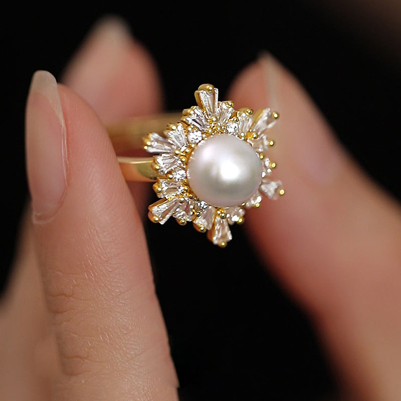 A gold ring with a central white pearl and surrounding clear baguette and round stones, shown worn on a person's fingers against a black background.