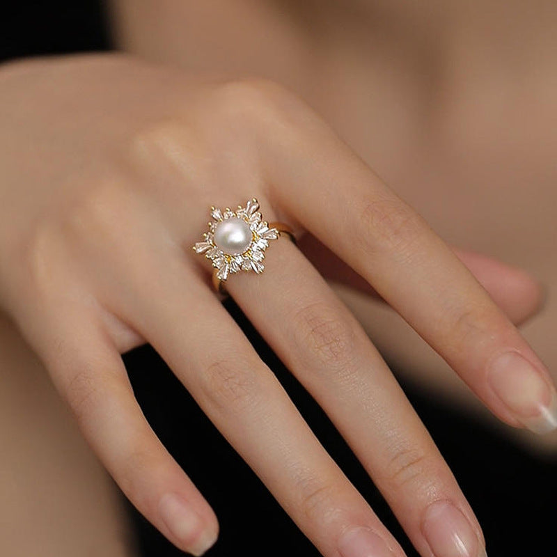 A close-up of a hand wearing a gold ring with a central white pearl and surrounding clear crystal-like accents.